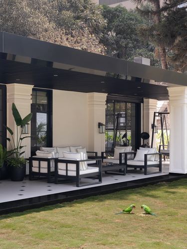 Covered veranda with black-framed white upholstered seating, cream columns, and two parrots on the lawn