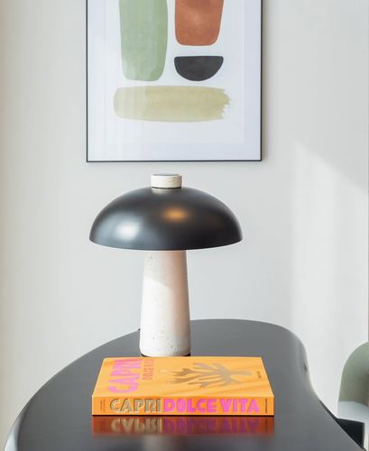 Black-domed mushroom lamp on a black table beside an orange Capri book, framed abstract art above.