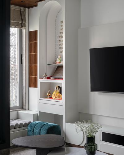 Arched pooja niche with deity idols beside a wall-mounted tv and teal ottoman