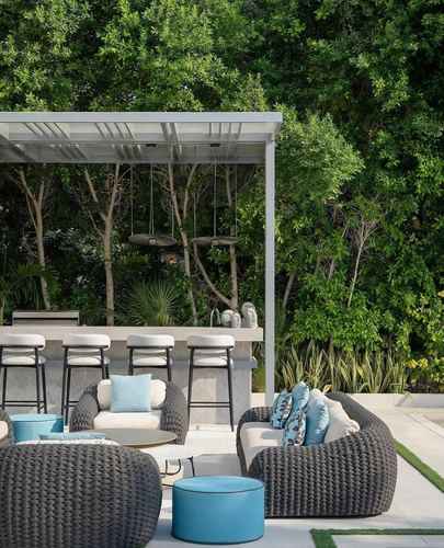 Outdoor patio lounge with grey woven chairs, blue pillows and pergola bar against greenery
