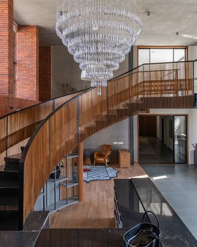 Curved wooden-slat staircase beneath large tiered crystal chandelier in double-height brick foyer