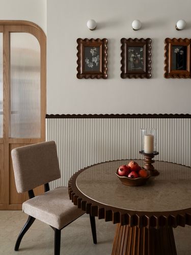 Round scalloped-edge dining table with pomegranates beneath three framed botanical prints on ribbed wainscot wall