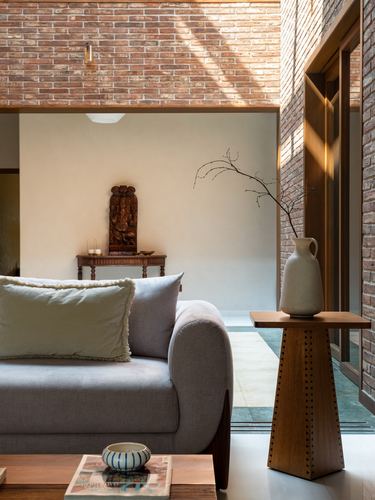 Grey sofa beside carved wooden side table with ceramic vase, exposed brick wall, sunlit interior niche