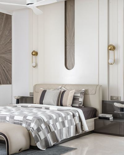Beige bedroom with quilted headboard, patterned throw, brass sconces, and wood inlay wall panel