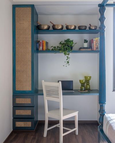 Teal built-in study nook with cane wardrobe panels, open shelves holding singing bowls and books, white chair at desk