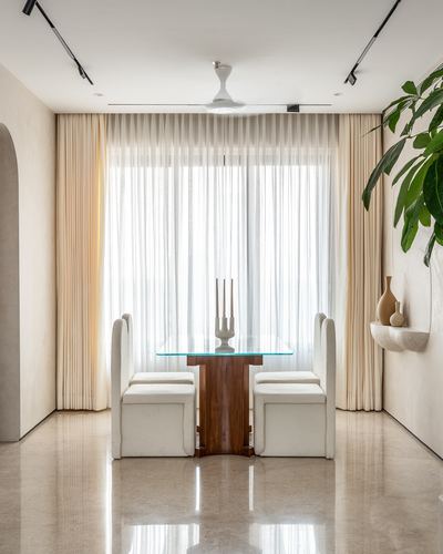 Dining nook with glass table on carved wood base, wingback chairs, sheer curtains, and shelf plant