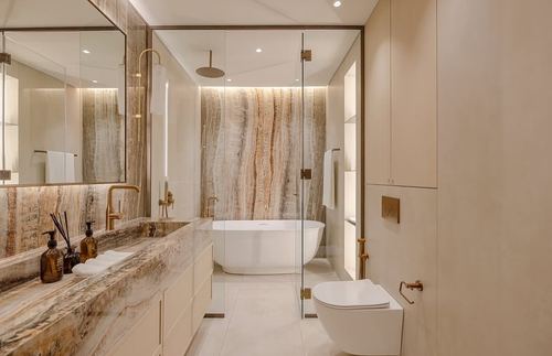Beige marble bathroom with freestanding tub, glass shower, brass fixtures and wood vanity