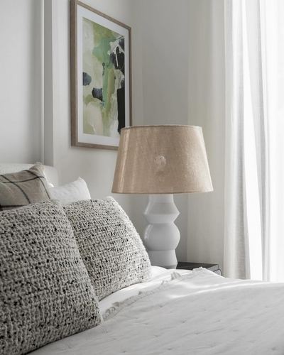 Bedside vignette with textured pillows, ceramic lamp, and framed abstract art on neutral wall