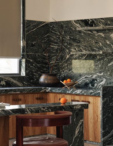 Corner of kitchen with dark veined marble backsplash and countertop, wood cabinetry, and a burgundy leather stool.