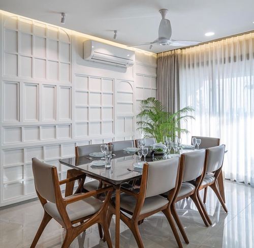 Dining room with white panelled wall, wooden chairs, glass-top table and sheer curtains