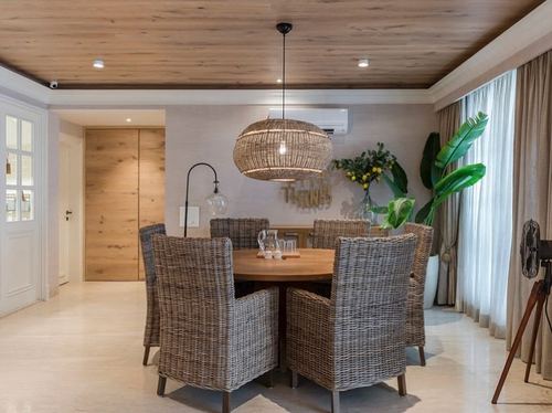 Round wood dining table with wicker chairs under a large rattan pendant light, wood-clad ceiling