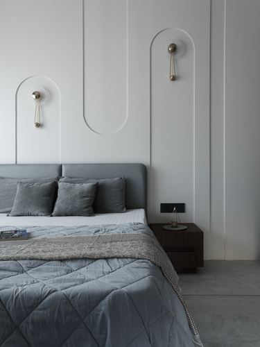 Grey upholstered bed against a white wall with arched recesses and sculptural wall sconces, dark wood nightstand beside it.