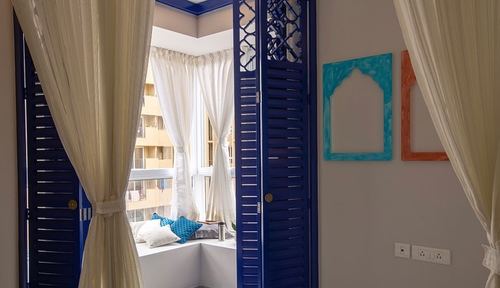 Blue carved shutters open to a curtained window seat with cushions and arched wall frames