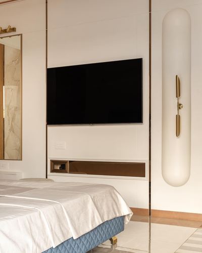 Bedroom wall with mounted TV, brass-trimmed panels, marble mirror and capsule sconce beside bed