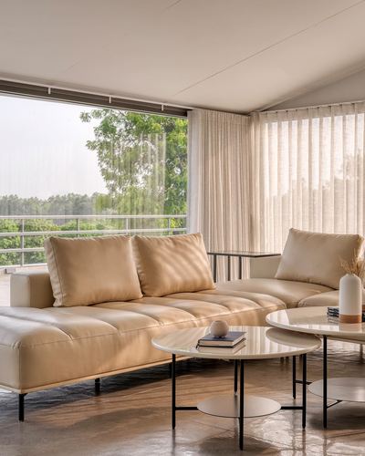 Beige tufted sofa with round marble coffee tables facing sheer-curtained balcony view of trees