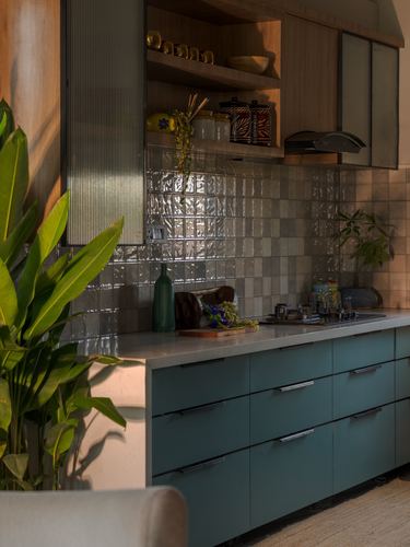 Kitchen corner with teal cabinetry, wood shelving, glazed tile backsplash, and large banana leaf plant.