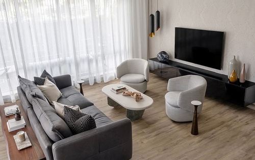 Living room with grey sofa, two cream swivel chairs, oval stone-base coffee table, black tv console, sheer curtains