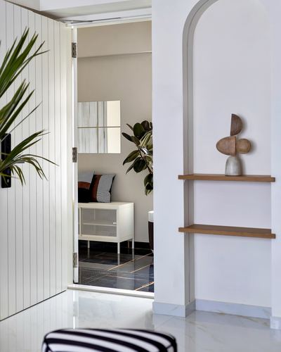 Arched niche with wooden shelves beside an open paneled door revealing a bedroom with mirror and plants.