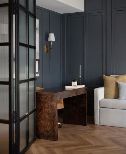 Home office with burl wood desk, dark panelled wall, brass sconce, and glass door in Dubai residence