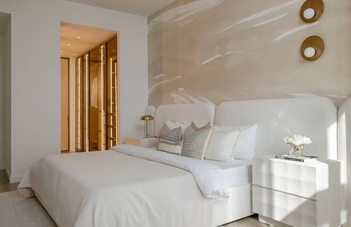 White upholstered bed against a beige marbled wall with brass disc sconces and glowing walk-in closet beyond