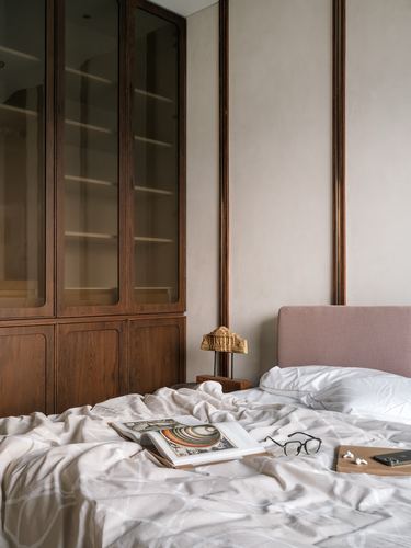 Bed with rumpled white linens beside a dark wood glass-front wardrobe and pink upholstered headboard