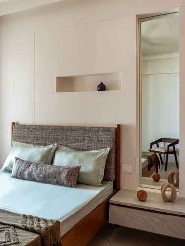 Minimal bedroom with tweed headboard, wall niche, and mirrored doorway reflecting a chair