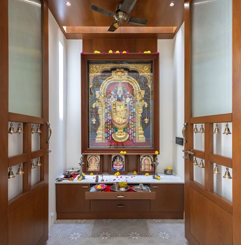Pooja room with wooden double doors, hanging bells, and large framed deity artwork above a marble counter