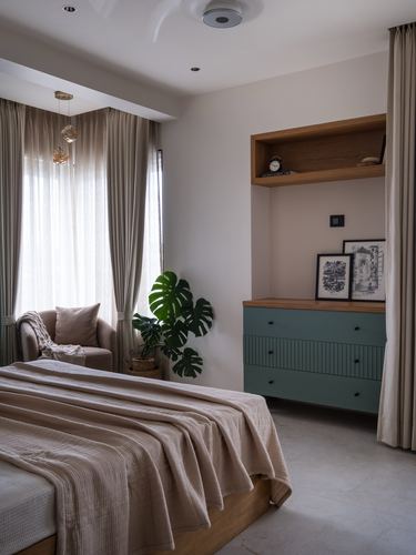 Bedroom corner with sage green dresser, wood niche shelf, armchair and monstera plant near bay window