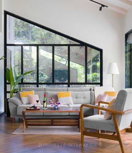 Sunlit living room with grey sectional, wooden coffee table, and angled black-framed glass wall facing greenery