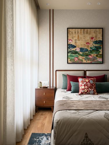 Bedroom with upholstered bed, teal and red cushions, lotus painting above, sheer curtains and wooden bedside table