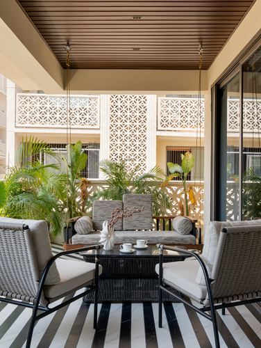 Balcony seating area with hanging swing, wicker chairs, and jali screen behind plants