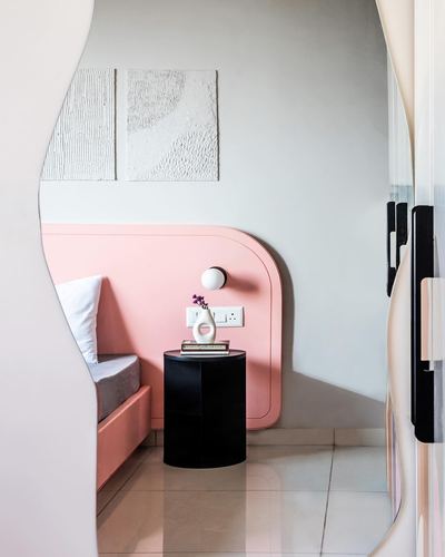 Curved pink headboard nook with black cylindrical side table and sconce, seen through a wavy wall opening.