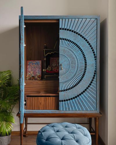 Blue carved wooden pooja cabinet open to reveal deity idols and framed prints, tufted stool nearby