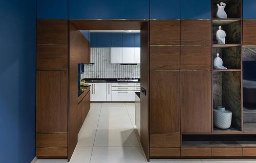 Walnut cabinetry wall with hidden door opening into a white and blue kitchen beyond