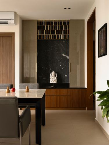Dining table in foreground with a black marble pooja niche and wooden cabinetry behind