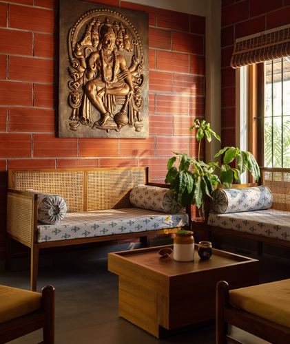 Cane daybeds with block-print bolsters facing a brass relief panel on a brick wall