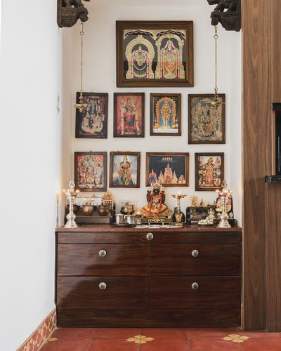 Pooja alcove with framed deity pictures above a dark wood cabinet on terracotta floor