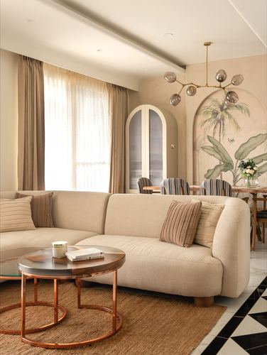 Beige sectional sofa with round nesting tables, arched mural, and chandelier in living room