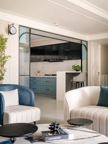 Living room with cream and teal armchairs opening to kitchen through arched glass partition