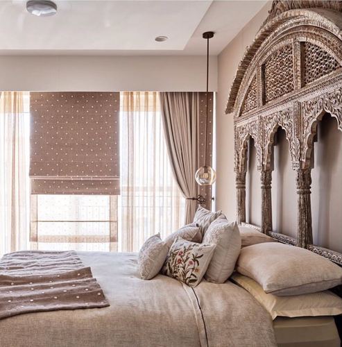 Bedroom with carved wooden arched headboard, layered neutral bedding, and polka dot roman blind by window