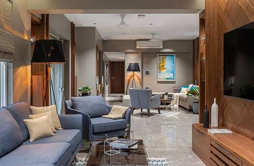 Open living room with blue sofas, marble flooring, wood paneling and tripod lamps, Pune apartment