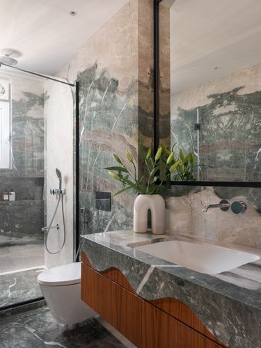 Marble-clad bathroom with wood vanity, wall-hung toilet, glass shower, and lily arrangement