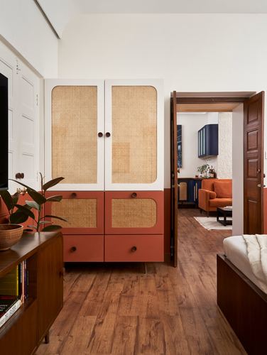 Terracotta and cane wardrobe beside open teakwood door revealing living room with orange sofa
