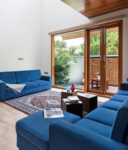 Blue velvet sectional sofas around a patterned rug facing a wood-framed glass door opening to a green courtyard