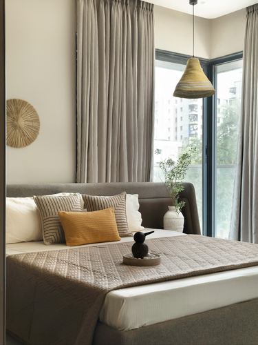 Beige upholstered bed with mustard cushions beneath a woven pendant light by corner window