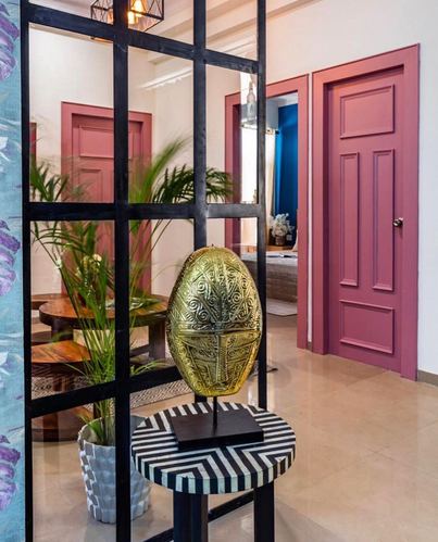 Foyer with black metal grid partition, brass mask sculpture on striped table, pink panelled doors beyond