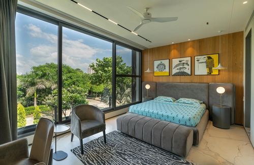 Modern bedroom with grey upholstered bed, wood-panelled wall, and floor-to-ceiling glass overlooking greenery