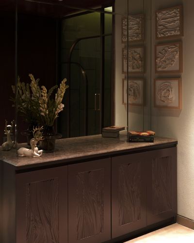 Dark wood console with carved cabinet doors and stone countertop beneath a mirrored wall with textured framed panels