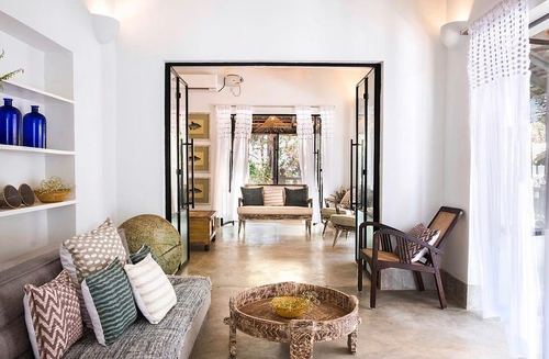 White living room with cane chair, carved wooden table, and view through glass doors to another seating area, Goa