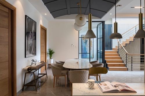Dining area with marble table, quilted chairs, brass pendants and curved grey ceiling, staircase beyond
